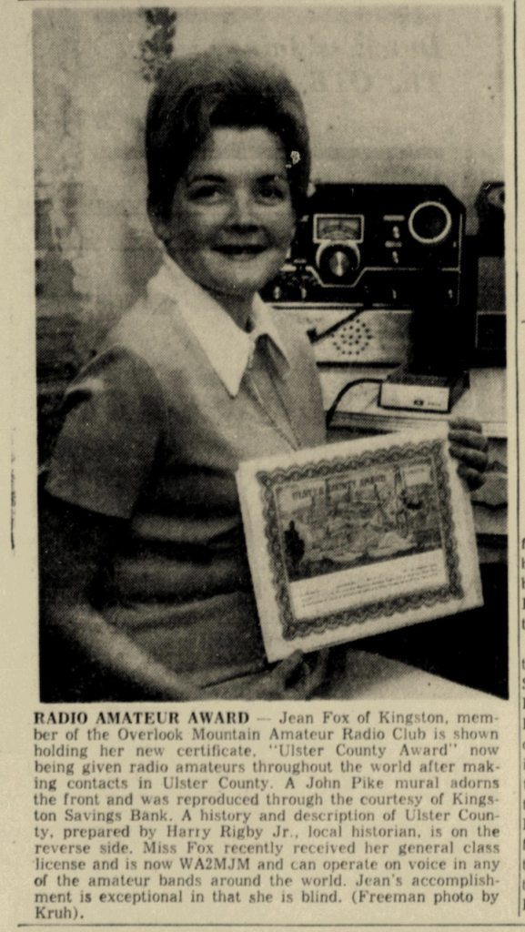 Jean sitting in front of her radio, holding the award.  Caption reads: RADIO AMATEUR AWARD. Jean Fox of Kingston, member of the Overlook Mountain Amateur Radio Club is shown holding her new certificate. "Ulster County Award" now being given radio amateurs throughout the world after making contacts in Ulster County. A John Pike mural adorns the front and was reproduced through the courtesy of Kingston Savings Bank. A history and description of Ulster County, prepared by Harry Rigby Jr., local historian, is on the reverse side. Miss Fox recently received her general class license and is now WA2MJM and can operate on voice in any of the amateur bands around the world. Jean's accomplishment is exceptional in that she is blind. (Freeman photo by Kruh).