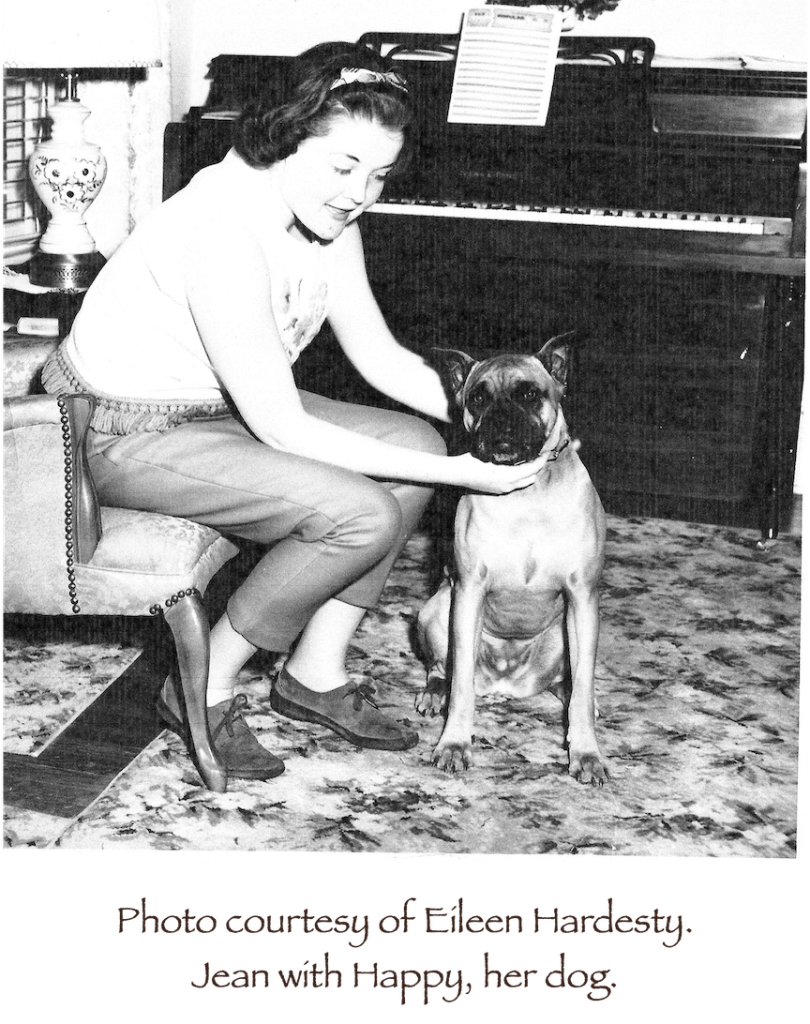 Jean in a chair by a piano, petting the dog.